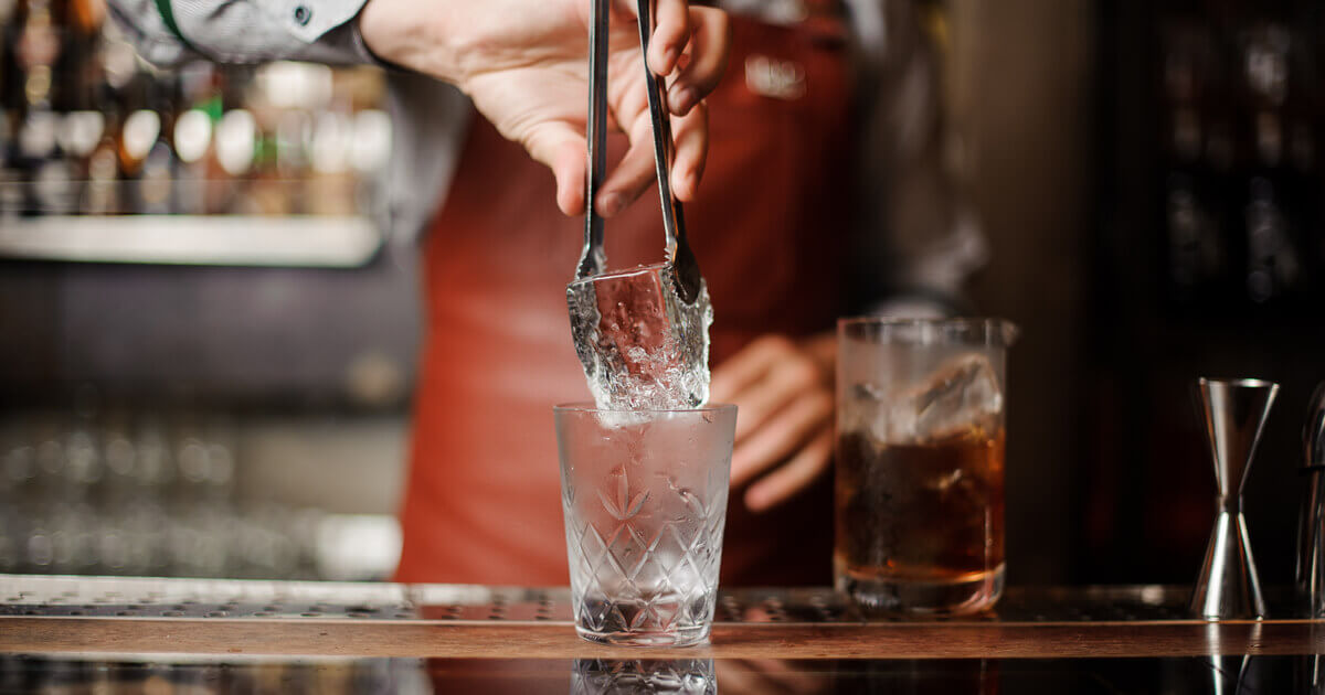 Ein großer Eiswürfel wird von einem Barkeeper in ein Glas gelegt